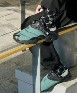 Person wearing Reebok x SOUTH2 WEST8 IPF Mule in green and black, styled with black graphic socks and urban outdoor pants, shown balancing on a metal rail in a city environment